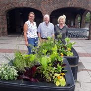 3 people standing behind two planters filled with vegetables ripe for harvest.