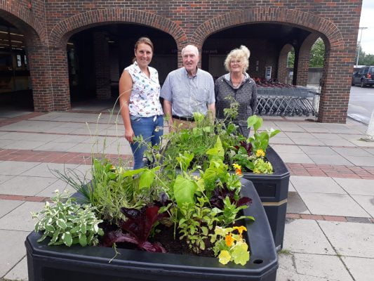 3 people standing behind two planters filled with vegetables ripe for harvest.