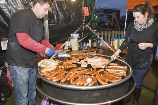 Two people cooking sausages on large round grill.