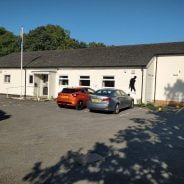 Outside of white single storey building with two cars parked in front.