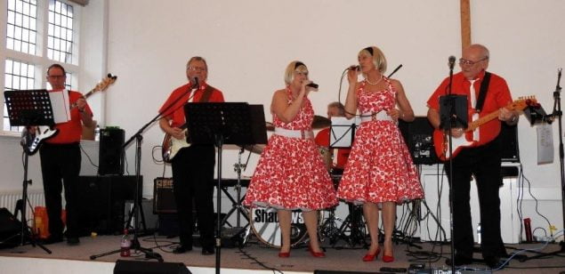 Band playing on a stage with two female singers in red dresses.