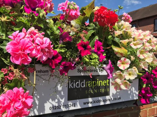 Close up of pink, purple and white flowers in a trough with a plaque on the container showing a sponsor's name.