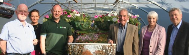 Group of six people standing in a poly tunnel. Hanging baskets behind.