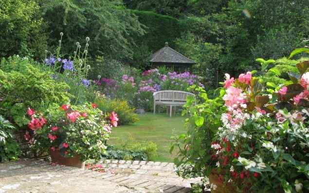 Large garden, flower beds to left and right, summer house and bench in background and patio in foreground.