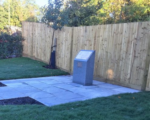 Wedge shaped grey stone war memorial standing on paved area in front of a new wooden fence. Grass and trees to side. 