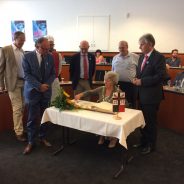 Mayor signs a book in a formal meeting room watched by a group of observers