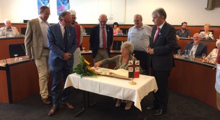 Mayor signs a book in a formal meeting room watched by a group of observers