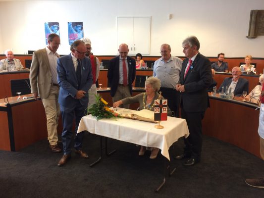 Mayor signs a book in a formal meeting room watched by a group of observers