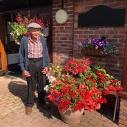 Elderly man standing next to container overflowing with red flowers and plants.