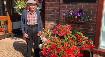 Elderly man standing next to container overflowing with red flowers and plants.