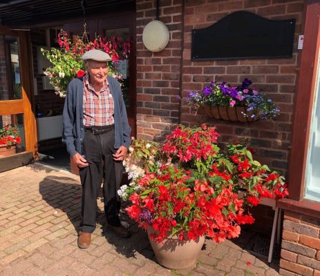 Elderly man standing next to container overflowing with red flowers and plants.