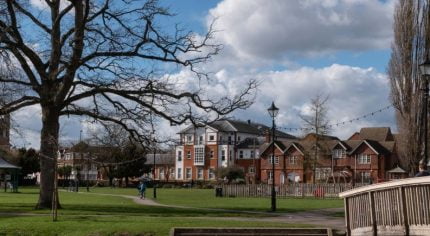 Winter scene of town centre park. Wooden bridge in foreground, trees, buildings and bandstand in background. Blue sky.