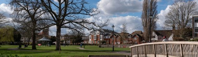 Winter scene of town centre park. Wooden bridge in foreground, trees, buildings and bandstand in background. Blue sky.