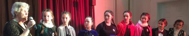 Female speaking into a microphone and eight children standing next to her in a row.