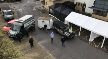 Aerial photo of four people in front of vehicles and a marquee.