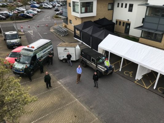 Aerial photo of four people in front of vehicles and a marquee.