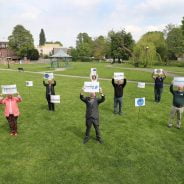 Group of people standing 2m apart on grass holding signs.