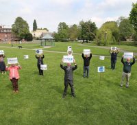 Group of people standing 2m apart on grass holding signs.