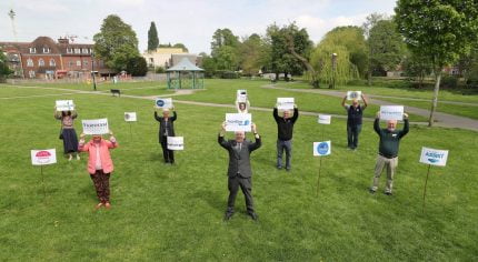 Group of people standing 2m apart on grass holding signs.