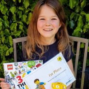 Girl sitting on bench in garden holding a certificate and lego prize.