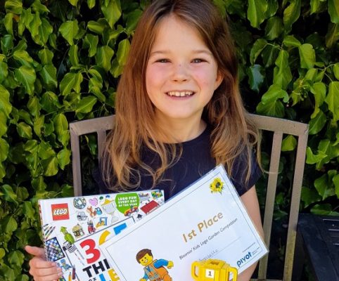 Girl sitting on bench in garden holding a certificate and lego prize.