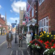 Armed Forces Day flag raising ceremony. Five people standing outside the town hall. Armed forces day flag and the Union flag are flying. Colourful flowers in a hop cart in the foreground.