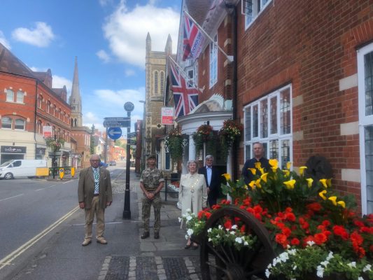 Armed Forces Day flag raising ceremony. Five people standing outside the town hall. Armed forces day flag and the Union flag are flying. Colourful flowers in a hop cart in the foreground.