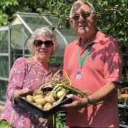 Man and woman holding a basket of potatoes and garlic. Greenhouse and trees in background