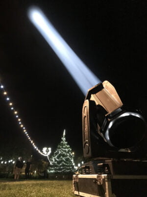Spotlight shines beam of light into sky. Christmas tree in background.