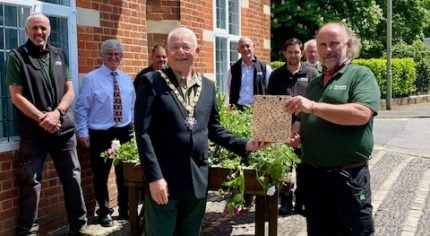 Mayor presents a gift to a male in a green uniform. Both are in front of a flower cart with people behind.