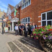 Group of people standing outside a red brick building with a hop cart filled with flowers in the foreground.