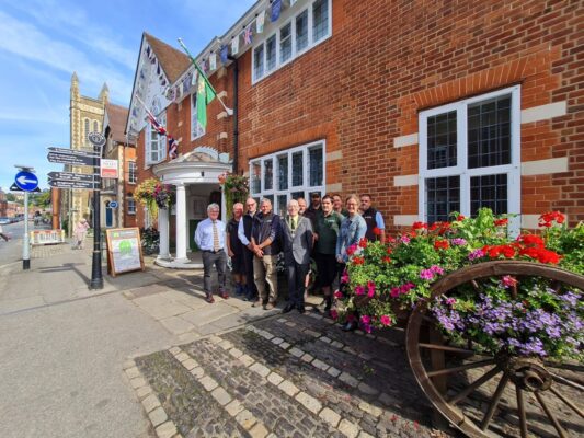Group of people standing outside a red brick building with a hop cart filled with flowers in the foreground.