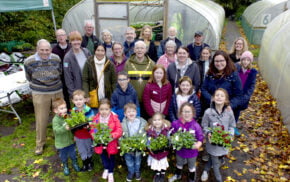 Group of children holding plants and a group of adults in front of a greenhouse.