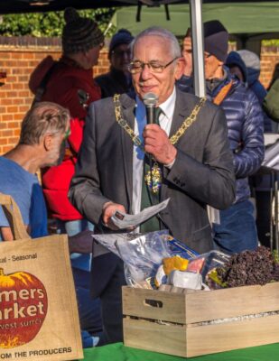Mayor holding a microphone with a box of fresh produce on table in front of him.