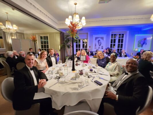 People at a black tie dinner. Sitting at a round table laid with white cloths.