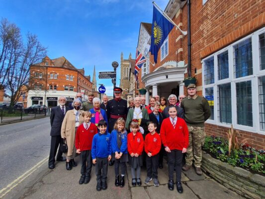Schoolchildren and adults stand under a flag