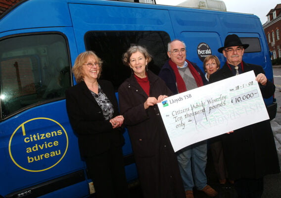 Five people holding a large cheque. Standing in front of a blue mini bus.