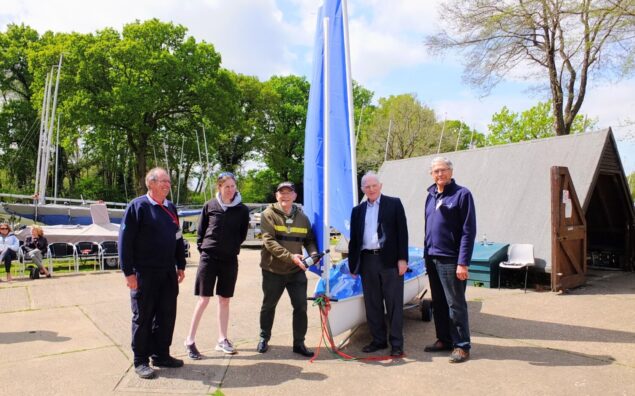 Five people standing in front of a sailing boat on land.