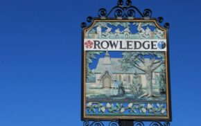 Ornate Rowledge Village Sign featuring a church, tree and men playing cricket against a blue sky.