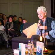 Female sitting on a grey sofa and talking to an audience.