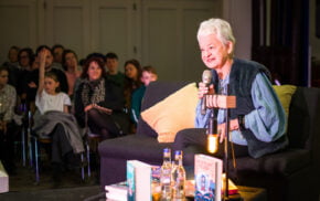 Female sitting on a grey sofa and talking to an audience.