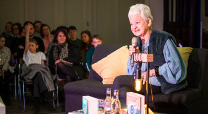 Female sitting on a grey sofa and talking to an audience.