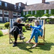 Two females sitting in deckchairs in a garden with a hotel in the background.