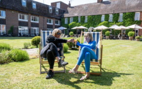 Two females sitting in deckchairs in a garden with a hotel in the background.