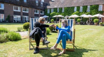 Two females sitting in deckchairs in a garden with a hotel in the background.