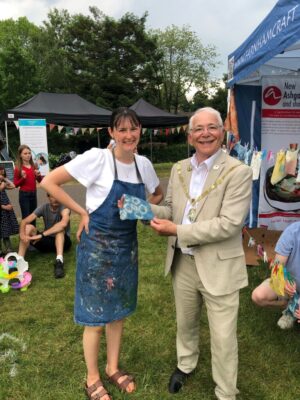 Lady in a paint splattered apron with Mayor holding a square of tie dyed material