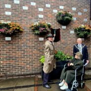 Two males and an older lady in a wheelchair unveiling a plaque on a brick wall.