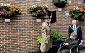 Two males and an older lady in a wheelchair unveiling a plaque on a brick wall.