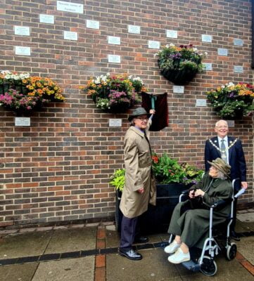 Two males and an older lady in a wheelchair unveiling a plaque on a brick wall.