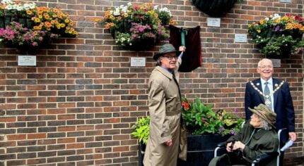 Two males and an older lady in a wheelchair unveiling a plaque on a brick wall.